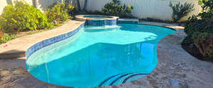 Freeform backyard swimming pool with blue waterline tile, built-in spa waterfall, and stone deck.